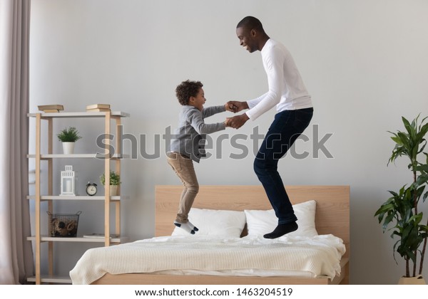 幸せな活動的な黒人の父親とかわいい小さな混血児の息子 がベッドマットレスで手の飛び跳びをし アフリカ系アメリカ人の子どもの子どもと一緒に楽しく寝室で笑う子どもを持つ 無料の家庭の父親 の写真素材 今すぐ編集