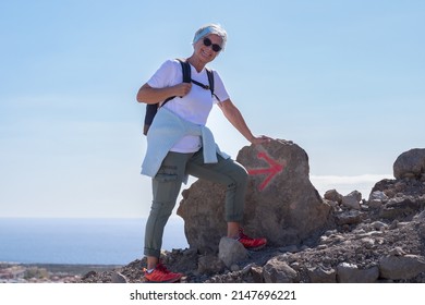 Happy Active Elderly Woman In Outdoor Hike Climbing In The Mountains With Backpack Following The Signs On The Trail Ahead. Horizon On The Water In The Background