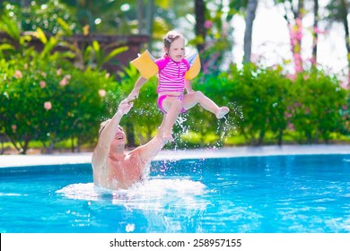 Happy Active Family, Young Father And His Cute Daughter, Adorable Toddler Girl, Playing In A Swimming Pool Jumping Into The Water Enjoying Summer Vacation In A Beautiful Tropical Island Resort