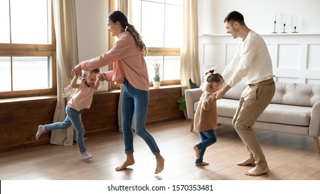 Happy active family young mom dad and cute little children daughters holding hands dancing together in living room interior, carefree funny small kids having fun jumping laughing with parents at home