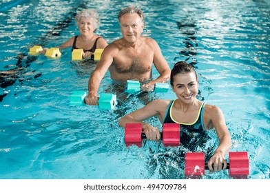 Happy active fitness mature man and senior woman doing exercise with aqua dumbbell in a swimming pool with instructor. Retired people doing aqua gym exercise with water dumbbell and looking at camera.