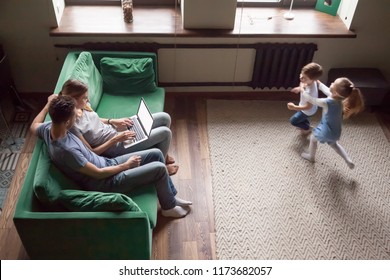 Happy Active Kids Running In Living Room Playing Game Having Fun Together While Parents Relaxing On Sofa Using Laptop, Family With Children Weekend Activities At Home Concept, Overhead Top View