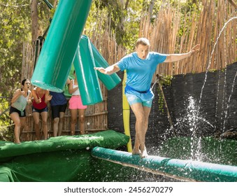 Happy active man overcoming obstacle course passing on soft log between hanging swinging bags over pool with water in outdoor amusement park on summer day .. - Powered by Shutterstock