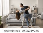 Happy active married couple dancing in stylish living room interior, enjoying activity at home together. Positive strong husband embracing wife in romantic social dance with passion