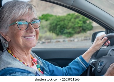 Happy Active Mature Adult Senior Woman Granny Driving The Car, Looking At Camera Smiling. Elderly Female Wearing Glasses Holding Wheel Enjoying Travel And Freedom With Her Automobile