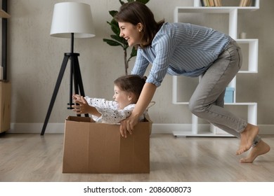 Happy active mom laughing, dragging box with screaming little daughter kid on floor at home. Mother and preschool child celebrating moving into new house, enjoying funny relocation party