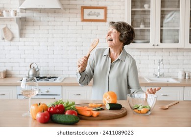 Happy active old senior elderly woman grandmother singing and using spoon as a microphone while cutting vegetables for salad, cooking at home kitchen - Powered by Shutterstock