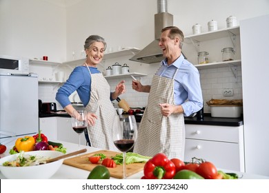 Happy Active Old Senior Mid Aged 60s Family Couple Enjoying Dancing And Cooking Healthy Meal Together At Home, Having Fun Preparing Romantic Dinner, Celebrating Valentines Day With Wine In Kitchen.