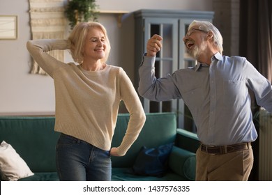 Happy active positive retired elder couple dancing together in living room, cheerful old senior husband and mature middle aged fit wife enjoying fun leisure activity laughing bonding moving at home