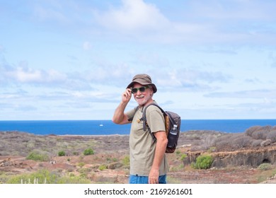 Happy Active Senior Man With Backpack And Hat While Hiking Outdoors Looking At Camera Smiling. Elderly Man Enjoying Retirement And Healthy Lifestyle. Blue Sea And Sky In The Background