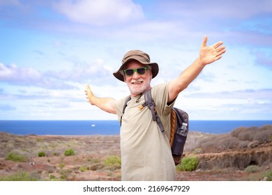 Happy Active Senior Man With Backpack And Hat While Hiking Outdoors Looking At Camera With Outstretched Arms. Blue Sea And Sky In The Background