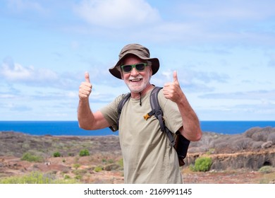 Happy Active Senior Man With Backpack And Hat While Hiking Outdoors Looking At Camera With Thumbs Up. Blue Sea And Sky In The Background