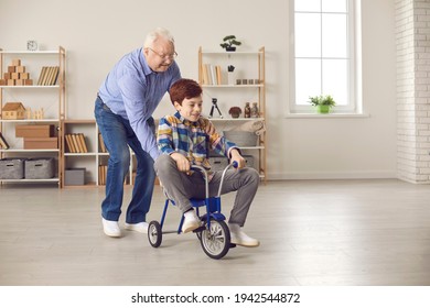 Happy Active Senior Man Playing Childish Games And Having Lots Of Fun With Grandson At Home. Little Kid Fooling Around With Energetic Grandpa, Enjoying Free Time And Riding Tricycle In The Living-room