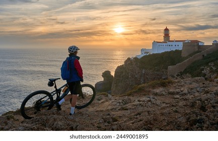 happy active senior woman cycling during moody golden hour near sunset at the the rock cliffs and lighthouse of Cabo Sao Vicente, the south-western spit of Europe at the atlantic coast of Portugal - Powered by Shutterstock