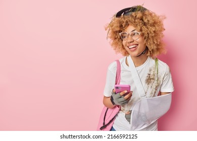 Happy Active Woman Cyclist Wears Sling On Arm Looks Cheerful Away Wears Dirty T Shirt And Helmet Uses Smartphone Concentrated Away Isolated Over Pink Background Empty Space For Your Advertisement