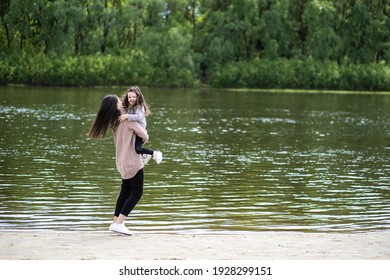 Happy active young mother and her little daughter hugging outdoor on spring scenic river beach on windy day. Family having fun outside together