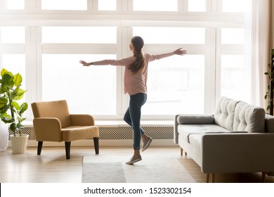 Happy Active Young Woman Dancing Alone In Modern Living Room Interior With Big Window Enjoying Cozy Weekend Lifestyle, Joyful Carefree Millennial Girl Listening Music Having Fun At Home Apartment