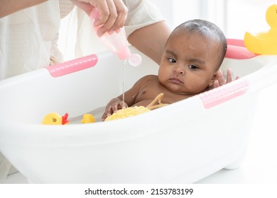 Happy Adorable African Newborn Baby Bathing In Bathtub. Mother Pouring Shower Gel On Sponge To Wash Her Little Daughter In Warm Water. Newborn Baby Cleanliness Care Concept