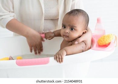 Happy Adorable African Newborn Baby Bathing In Bathtub. Mother Washing Her Little Daughter In Warm Water With Yellow Sponge And Shower Gel. Newborn Baby Cleanliness Care Concept