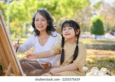 Happy and adorable Asian little granddaughter painting watercolor on canvas with her grandmother in the city park on the weekend. happy family time concept - Powered by Shutterstock