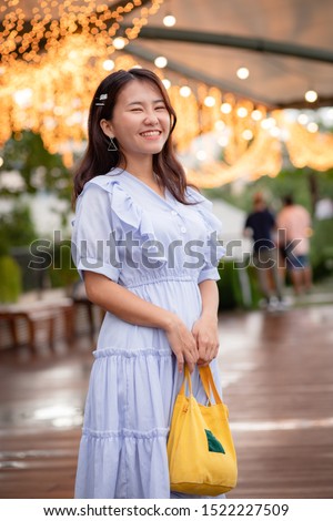Happy adorable Asian woman in light blue dress carrying a small yellow tote bag who standing on riverside with warm light decoration in background