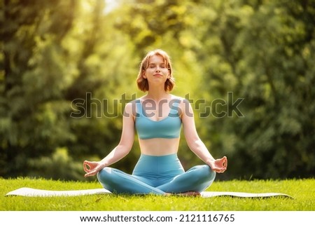Happy adorable athletic girl performing yoga sitting on mat in lotus position outdoors against background of green trees in city park, sporty female  during meditation practice