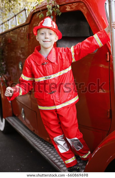 Happy Adorable Child Boy\
with Fireman Hat Playing Outside standing on old shiny vintage red\
fire truck. Dreaming of future profession. Fire safety, Life\
Protection lessons