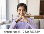 Happy adorable cute Indian kid girl showing hand heart shaped sign, symbol of love, family, care, support, charity, looking at camera with toothy smile, enjoying childhood