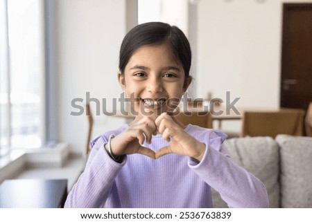 Happy adorable cute Indian kid girl showing hand heart shaped sign, symbol of love, family, care, support, charity, looking at camera with toothy smile, enjoying childhood