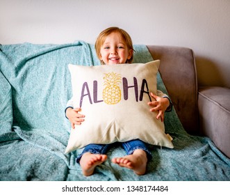 Happy, Adorable, Cute Toddler Boy Wearing Blue Jeans And White Sweater Holding Aloha Pillow Sits On Couch With Blue Throw Blanket.