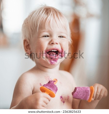 Happy adorable infant baby boy child smiling while eating two frozen fruit popsicle ice creams in simmer.