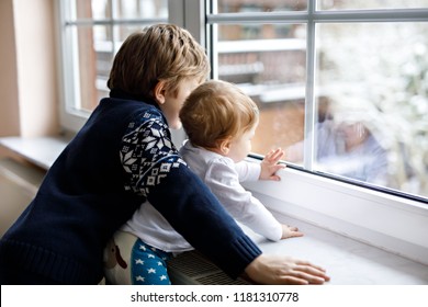 Happy Adorable Kid Boy And Cute Baby Girl Sitting Near Window And Looking Outside On Snow On Christmas Day Or Morning