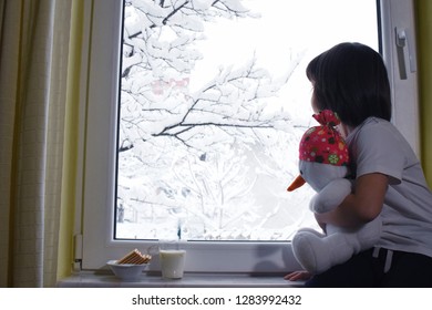 Happy adorable kid boy sitting near window and fascinated looking outside on snowfall on Christmas day. Child eat cookies and drink milk near window. Child hugging toy Snowman in front of window