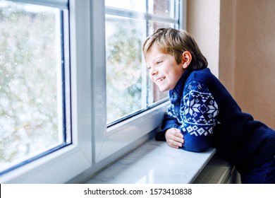 Happy Adorable Kid Boy Sitting Near Window And Looking Outside On Snow On Christmas Day Or Morning. Smiling Healthy Child Fascinated Observing Snowfall And Big Snowflakes