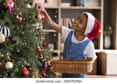 Happy adorable little 7s African American kid girl in red hat decorating Christmas tree with beautiful toys, taking baubles from basket, preparing for winter New Year holidays alone at home. - Powered by Shutterstock