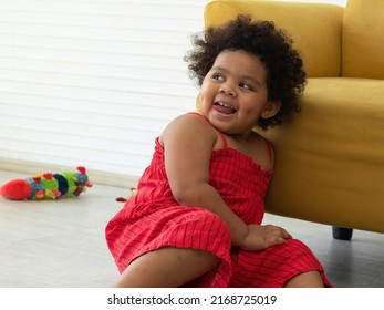 Happy Adorable Little African Girl With Afro Hair Wearing Red Jumpsuit Laughing With Happiness On The Floor Of Living Room At Home.