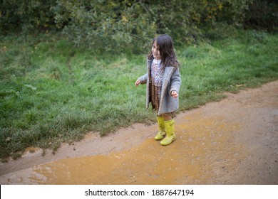 Happy Adorable Little Girl With Muddy Clothes Smiling And Looking At Puddle Before Jumping In It. Young Child Having Fun Jumping In Puddles. Kids Playing Outside