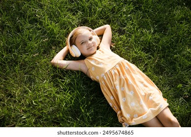 Happy  adorable smiling child girl in headphones listens to music lying on the green grass in the garden in summer. Happy ghild having fun outdoors. Generation Alpha.   copy space. favorite music. - Powered by Shutterstock