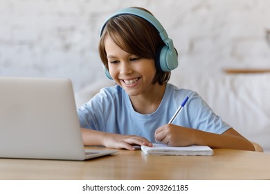 Happy adorable teenage kid boy in wireless headphones looking at laptop screen, listening educational online lecture, enjoying studying distantly at home, writing notes in copybook, e-learning concept