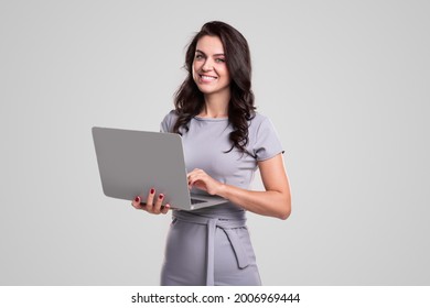Happy adult business lady in elegant dress looking at camera while working on laptop against gray background