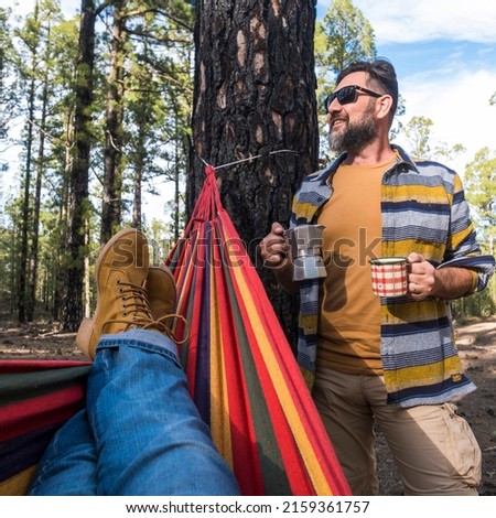 Happy adult caucasian couple enjoy the nature outdoor leisure camping activity - woman legs pov on hammock and man standing with coffee - environment and nature vacation