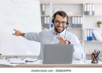Happy adult caucasian guy teacher in glasses shows at board, explain english rules, looks at laptop webcam, talk with student in home office interior. Seminar, webinar, e-learning and lesson, covid-19