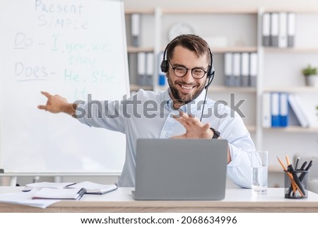 Happy adult caucasian guy teacher in glasses shows at board, explain english rules, looks at laptop webcam, talk with student in home office interior. Seminar, webinar, e-learning and lesson, covid-19