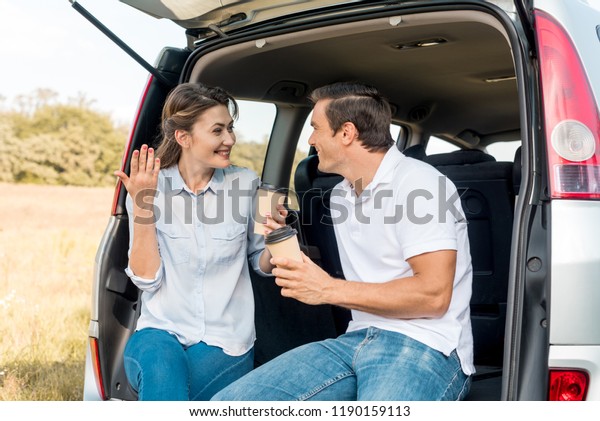 happy adult couple with paper cups of\
coffee sitting in car trunk while travelling by\
car