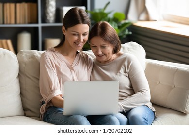 Happy Adult Daughter And Mature Mother Using Laptop Together, Sitting On Couch At Home, Smiling Young Woman And Older Mom Surfing Internet Or Shopping Online, Looking At Computer Screen