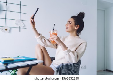 Happy Adult Female In Casual Clothes Focusing On Screen And Taking Selfie With Smartphone While Sitting In Armchair At Desk And Holding Take Away Glass With Orange Drink And Straw In Light Apartment 