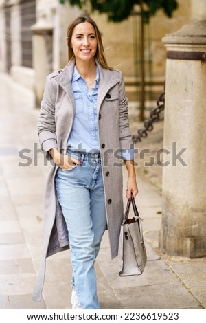 Happy adult female in casual clothes and gray coat with stylish bag holding hand in pocket and smiling while strolling on sidewalk on city street