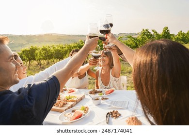 Happy adult friends having fun drinking red wine and eating together with vineyard in background - Multiracial people doing appetizer at summer time in countryside resort - Main focus on hands - Powered by Shutterstock