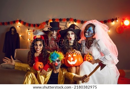 Happy adult friends in spooky costumes posing for funny group photo. Cheerful young people dressed as crazy witch, devil, vampire, dead bride and clown with axe having fun together at Halloween party