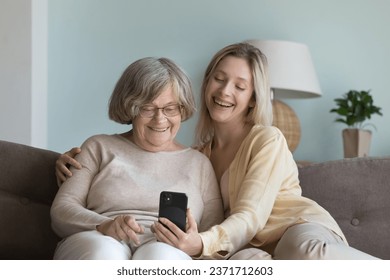 Happy adult granddaughter girl and cheerful eldest grandmother speaking on video conference call, holding mobile phone, enjoying online Internet communication, relaxing on home couch - Powered by Shutterstock